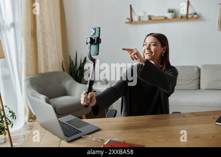 Eine junge Frau filmt sich mit einem Handy auf einem Selfie-Stick, während sie an ihrem Schreibtisch sitzt. Stockfoto