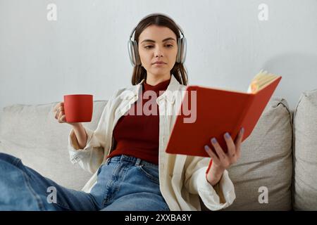 Eine junge Frau in lässiger Kleidung genießt einen Moment der Entspannung in ihrem Wohnzimmer und hört Musik, während sie ein Buch liest. Stockfoto