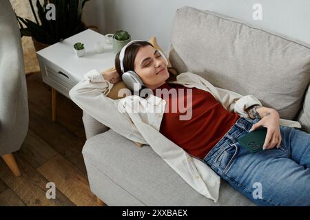 Eine junge Frau in lässiger Kleidung liegt auf einer Couch, trägt Kopfhörer und lächelt, während sie zu Hause ausruht Stockfoto