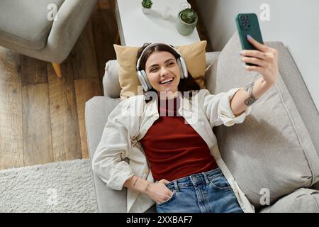 Eine junge Frau in lässiger Kleidung entspannt sich auf einer Couch, Kopfhörer an, macht ein Selfie auf ihrem Handy. Stockfoto