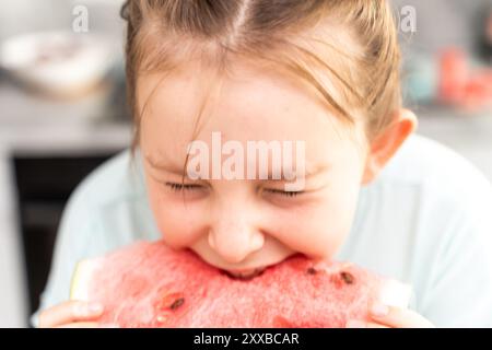 Ein kleines Teenager-Mädchen hält ein Stück Wassermelone in den Händen und beißt es mit geschlossenen Augen hinein und genießt den Geschmack. Vor dem Hintergrund von t Stockfoto