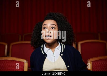 Eine junge Frau in einer dunkelblauen Jacke sitzt in einem Kino und sieht mit einem schockierten Ausdruck den Bildschirm. Stockfoto