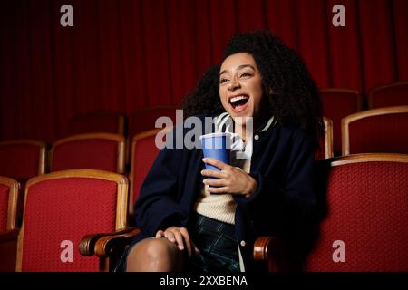 Eine junge Frau in stilvoller Freizeitkleidung lacht herzhaft, während sie im Kino einen Film ansieht und eine blaue Tasse in einer Hand hält. Stockfoto