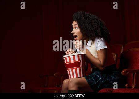 Eine junge Frau, lässig gekleidet, sitzt in einem Kinositz und sieht sich einen Film mit aufgeregtem Ausdruck an und hält einen Eimer Popcorn. Stockfoto