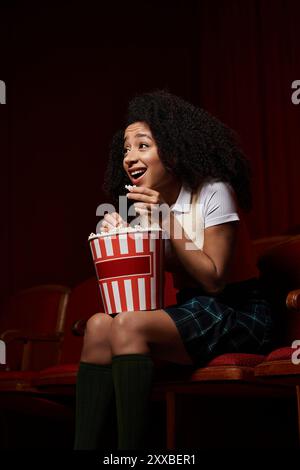 Eine schwarze Frau mit lockigen Haaren in einem Kinosaal reagiert aufgeregt, während sie Popcorn hält. Stockfoto