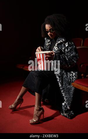 Eine modische Afroamerikanerin in einem schicken Pelzmantel sitzt in einem Kino, vertieft in den Film und genießt einen Eimer Popcorn. Stockfoto