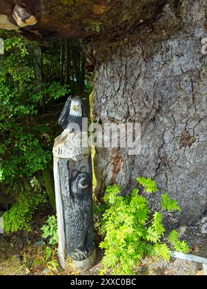 Ein kleines hölzernes Totem, das einen Bären, Fisch und Weißkopfseeadler im natürlichen Stil darstellt, steht am Fuß eines großen Baumes Stockfoto