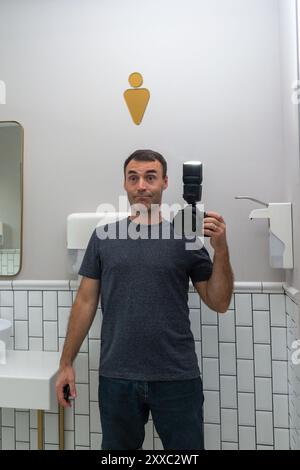 Ein Mann mittleren Alters mit einem dummen Lächeln im Gesicht macht ein Selfie im Spiegel einer öffentlichen Toilette mit einer digitalen Spiegelreflexkamera mit Blitz Stockfoto