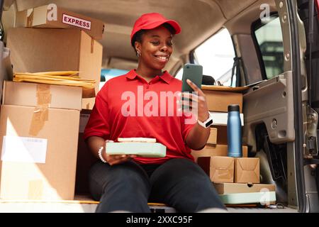 Lächelnder Mitarbeiter, der das Paket hält, während er das Smartphone im Lieferfahrzeug ansieht, umgeben von lächelnden Kisten und in roter Uniform Stockfoto