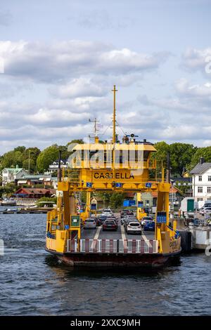 Eine gelbe Fähre namens Castella, die an einem Dock ankommt, mit Autos an Bord. Die Szene zeigt einen ruhigen Wasserweg umgeben von grünen Bäumen und Gebäuden in der Th Stockfoto