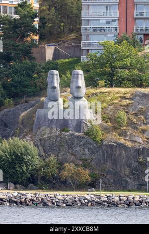 Zwei große Steinstatuen, die an die Osterinsel Moai erinnern, auf einem felsigen Hügel in der Nähe einer Uferpromenade gelegen, mit modernen Gebäuden im Hintergrund und gre Stockfoto