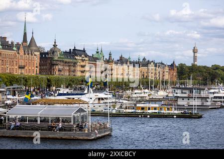 Ein malerischer Blick auf eine Uferpromenade in Stockholm, Schweden, mit historischen Gebäuden, Booten und schwedischen Flaggen. Der Himmel ist teilweise bewölkt, was den pi verstärkt Stockfoto