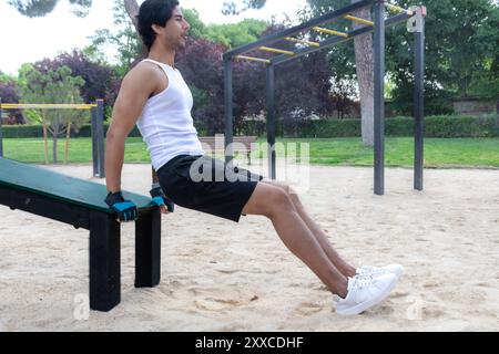 Ein Mann in einem weißen Tanktop sitzt auf einer Bank im Park. Die Bank befindet sich neben einer Metallkonstruktion mit einem gelben Stab Stockfoto