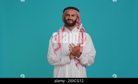 Arabischer Erwachsener in traditioneller Kleidung, klatscht im Studio die Hände, gratuliert und jubelt der Kamera mit selbstbewusstem Ausdruck an. Muslimischer Kerl zeigt Unterstützung und applaudiert Menschen. Stockfoto