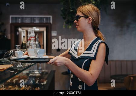 Cafe Kellnerin mit Tablett mit Kaffeetassen in stilvoller Uniform und Sonnenbrille in gemütlichem Café-Ambiente Stockfoto