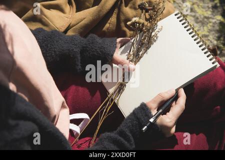 Nahaufnahme der Hände eines Mädchens, das ein leeres Notizbuch hält. Ein trockener Strauß Kräuter in der Hand und ein Bleistift. Reise-Designer-Künstler Stockfoto