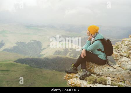 Junge Frau mit Rucksack, die pensiver am Rande eines Felsens sitzt und mit Wolken in den Himmel blickt Stockfoto