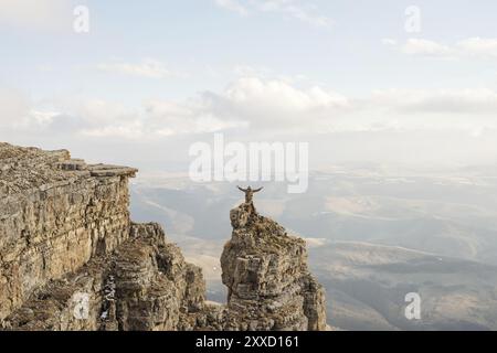 Ein glücklicher Mann mit erhobenen Händen steht auf einem separat stehenden Felsen, der über den Wolken vor dem Hintergrund von Tälern, Hügeln, Sonnen steht Stockfoto