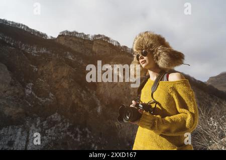 Eine Fotografin in Sonnenbrille und einem großen Pelzhut und einem gelben Strickpullover steht vor dem Hintergrund hoher Felsen in der Schlucht mit einem Camer Stockfoto