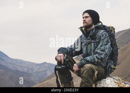 Müder bärtiger Hipster mit Fernglas in den Händen sitzt auf einem Stein zwischen den kaukasischen Bergen und blickt in die Ferne Stockfoto