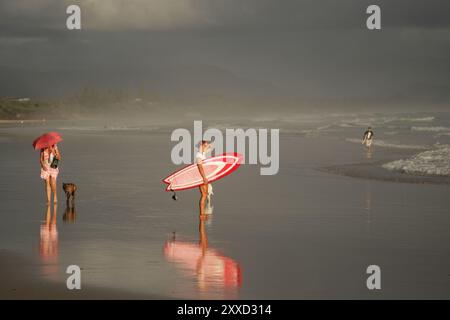 Junge Surfer mit rotem Surfbrett blicken am frühen Morgen auf das Meer am Strand in Byron Bay, Australien. Neben ihr eine Frau mit einem roten Regenschirm Stockfoto