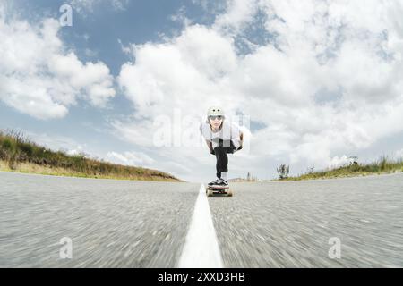 Der Typ mit Helm und Sonnenbrille fährt sehr schnell auf der Asphaltstraße in einem speziellen Sportständer sein Longboard Stockfoto