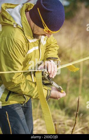 Ein Mann passt das Slacklining der Ausrüstung an, bevor er verrückte Slackliner-Tricks ausführt und in einem Park in der Natur auf einem Slacklin läuft Stockfoto