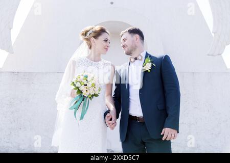 Porträt eines wunderbaren Paares, das an einem Hochzeitstag mit einem Blumenstrauß in der Hand vor dem Hintergrund eines orthodoxen christlichen Denkmals mit Engeln verbrachte. Stockfoto