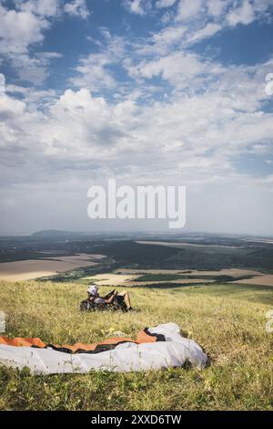 Ein professioneller Gleitschirm in voller Ausrüstung und ein Helm liegt und ruht auf dem Gras hoch in den Bergen und schaut auf die Wolken Stockfoto