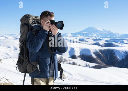 Porträt eines bärtigen Hipsters, eines Fotografen mit Rucksack und Sonnenbrille macht Fotos seiner DSLR vor dem Hintergrund des schneebedeckten Kaukasi Stockfoto