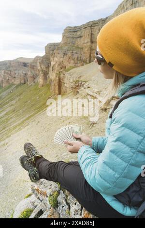 Ein Reisender mit Hut und Sonnenbrille hält hundert Dollar-Scheine in den Händen eines Ventilators vor dem Hintergrund von Felsen auf der Natur. Reisekosten Stockfoto