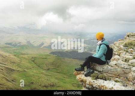 Junge Frau mit Rucksack, die pensiver am Rande eines Felsens sitzt und mit Wolken in den Himmel blickt Stockfoto