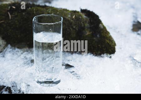 Transparentes Glasglas mit Trinkwasser im Winter steht auf einer Eiskruste vor dem Hintergrund eines sauberen, frostigen Flussufers. Der Tiefenbereich Stockfoto
