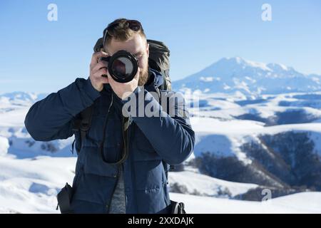 Porträt eines bärtigen Hipsters, eines Fotografen mit Rucksack und Sonnenbrille macht Fotos seiner DSLR vor dem Hintergrund des schneebedeckten Kaukasi Stockfoto