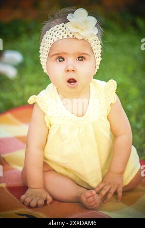 Cute Baby Mädchen in gelben Band und Kleid sitzt in sonnigen Hinterhof. 8 Monate altes Kleinkind im Freien. Entwicklungskonzept Stockfoto