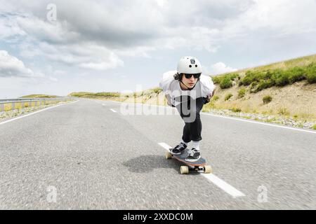 Der Typ mit Helm und Sonnenbrille fährt sehr schnell auf der Asphaltstraße in einem speziellen Sportständer sein Longboard Stockfoto