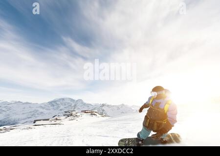 Ein Snowboarder in Skimaske und Rucksack fährt auf einer schneebedeckten Piste und hinterlässt Schneepulver vor dem blauen Himmel und der untergehenden Sonne. Stockfoto