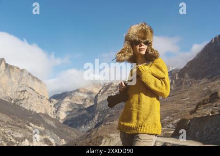 Eine Fotografin in Sonnenbrille und einem großen Pelzhut und einem gelben Strickpullover steht vor dem Hintergrund hoher Felsen in der Schlucht mit einem Camer Stockfoto