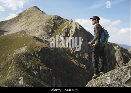 Ein bärtiger Mann mit Sonnenbrille und einer Mütze mit Rucksack steht auf einem Felsen und blickt in ein felsiges Tal hoch in den Bergen. Das Konzept der Tour Stockfoto