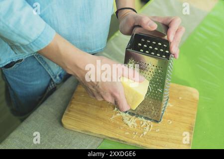 Nahaufnahme der Hände einer Frau reiben einen Parmesankäse auf einer Metallreibe auf einem grünen Tisch Stockfoto