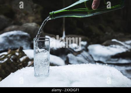 Mineralwasser wird aus einer glasgrünen Flasche in ein klares Glas-Becherglas gegossen. Ein Glas steht im Schnee. Vor dem Hintergrund eines Winters Stockfoto