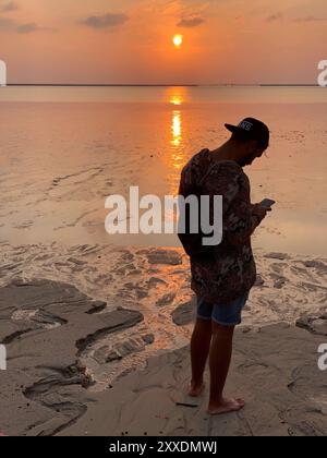 Reisende barfuß an einem Strand in Langkawi, Malaysia, mit Blick auf das Telefon bei Sonnenuntergang bei Ebbe und der Reflexion des Sandes im Wasser Stockfoto