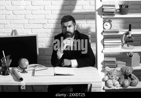 Porträt eines männlichen Lehrers aus der Vorschule oder Grundschule, der am Schreibtisch in der Klasse arbeitet. Reifer Mann sitzt entspannt in einem Klassenzimmer Blick auf die Stockfoto