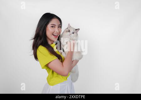 Lächelnde junge asiatische Frau im gelben Hemd hält eine weiße Ragdoll-Katze über isoliertem weißen Hintergrund. Stockfoto