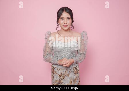 Schöne junge indonesische Frau, die modernes Kebaya Kleid trägt, steht und lächelt vor der Kamera über isoliertem rosa Hintergrund. Stockfoto