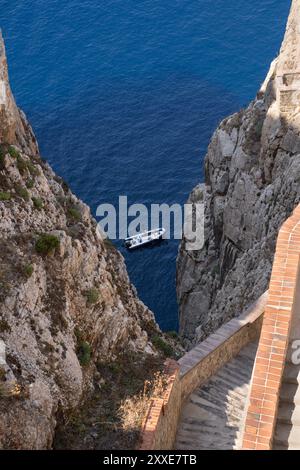 Ein kleines Boot fährt durch das tiefblaue Meer zwischen den zerklüfteten Klippen der Neptun-Grotte, die von der Steintreppe, die nach Sardin führt, erfasst wurde Stockfoto