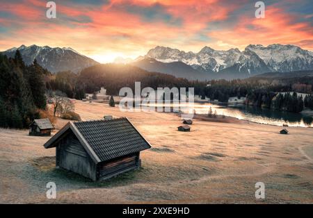 Sonnenaufgangslandschaft an einem See, mit einer Hütte im Vordergrund und Bergen im Hintergrund, gefrorener Wiese und bunten Wolken am Himmel Stockfoto