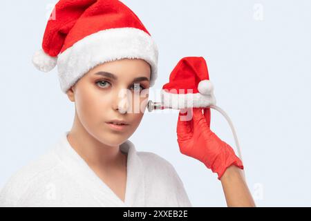 Der Kosmetiker macht die Prozedur Mikrodermabrasion auf dem Gesicht einer schönen Frau mit einem Weihnachtsmann-Hut. Neujahrs- und Kosmetologiekonzept. Stockfoto