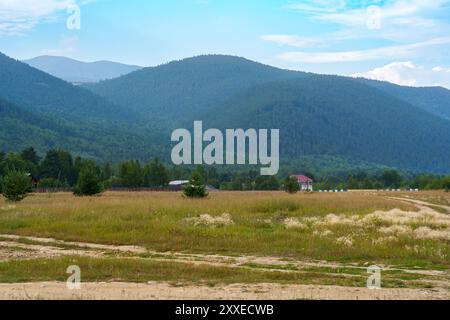 Eine unbefestigte Straße schlängelt sich durch ein grasbewachsenes Feld in Richtung eines bunten Zauns, der vor dem Hintergrund nebeliger Berge unter einem teilweise bewölkten Himmel liegt und ein VI erzeugt Stockfoto