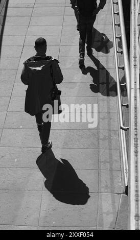 Ein beeindruckendes Schwarz-Weiß-Hochwinkelbild, das zwei Fußgänger zeigt, die über einen gepflasterten städtischen Platz gehen, ihre langen Schatten sich dehnend Stockfoto